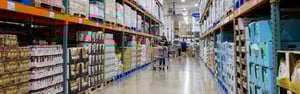 Customers walking through wide aisles of a large wholesale club, filled with bulk inventory on high shelves.