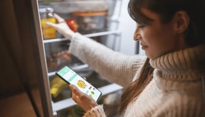 In her home, a woman combs through her refrigerator while ordering groceries online using her phone. 